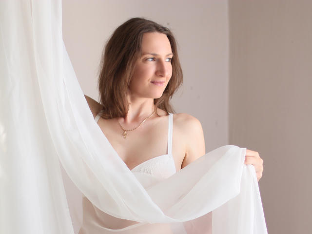AngelicaShackley with brown hair, white lingerie, holding a sheer white curtain, smiling, wearing a gold necklace with a cross, soft natural light. Her tits are visible.