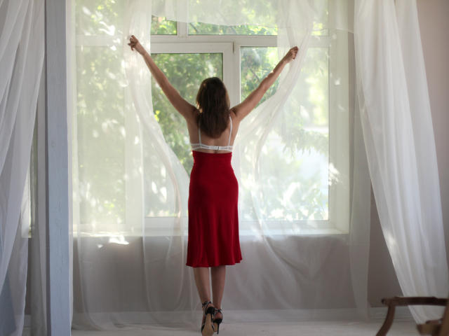 AngelicaShackley stands with arms raised, holding white curtains, wearing a red skirt and black heels, back to the camera, in a sunlit room, showing her ass.