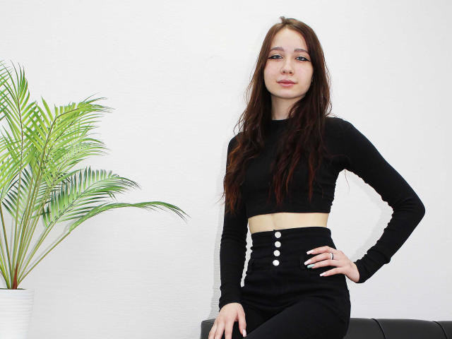 AdrianeWalter with long brown hair, wearing a black crop top and high-waisted black skirt with white buttons, standing with hand on hip, next to a green plant.