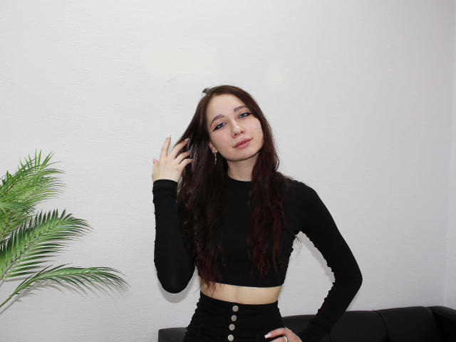 AdrianeWalter with long brown hair, wearing a black crop top and high-waisted black pants, hand in hair, standing next to a green plant, looking at camera.