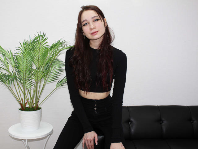 AdrianeWalter with long brown hair, black long-sleeve crop top, and black button-up pants, sitting on black leather couch, potted green plant to left, white wall background.