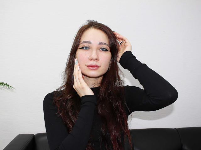 AdrianeWalter with long, dark brown hair, wearing a black long-sleeve shirt, has a hand in her hair and the other touching her cheek, sitting on a black couch.