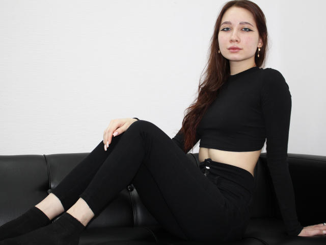 AdrianeWalter with long brown hair, black crop top, black pants, and black socks, sitting on black leather couch, legs crossed, hands on couch, looking at camera, white background.