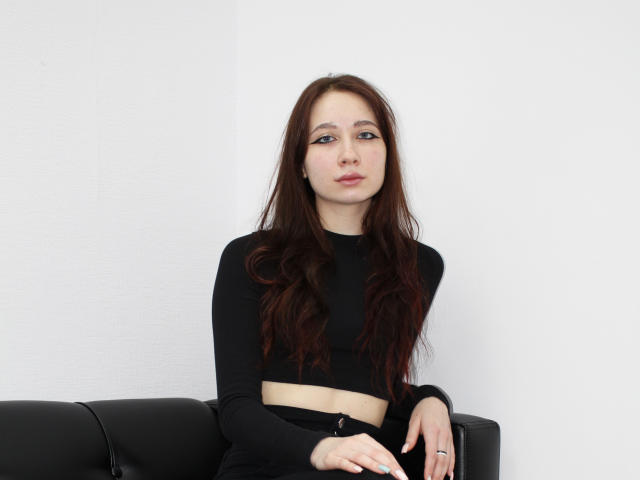 AdrianeWalter has long brown hair, wearing a black crop top and black skirt, sitting on a black couch, with a neutral expression, hands on her lap.