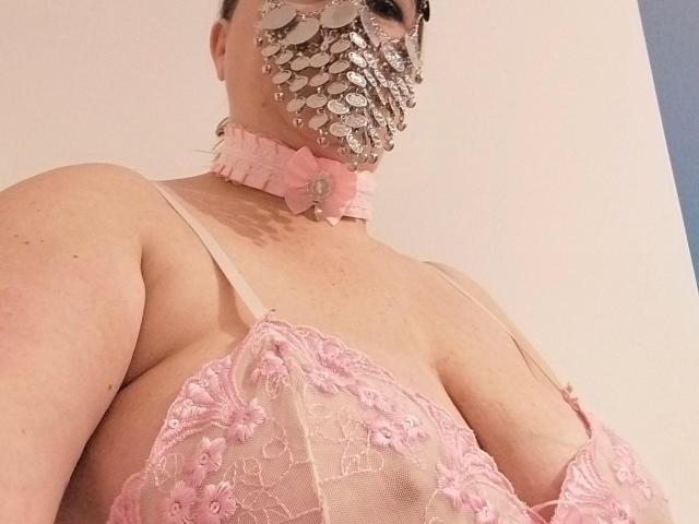 LiliChattePoilue with big tits, wearing a pink lace bra and a silver, jeweled face mask with a pink choker, shot from a low angle.