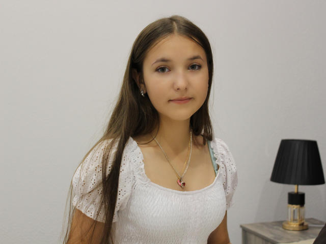 AngelikaFulton with long brown hair, wearing a white lace top, small tits, and a necklace with a red heart, standing next to a black lamp on a wooden table.