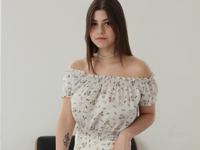 Mary Gibbs, con i capelli castani, indossa una camicetta bianca a fiori con spalline scoperte, una collana choker, ha un leggero sorriso, un tatuaggio sul braccio destro, è in piedi, guarda la fotocamera, sfondo bianco. Ha il seno, un piccolo tatuaggio, un aspetto casual..
