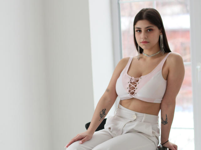 Mary Gibbs mit einem hellrosa Sport-BH, der ihre Brüste zeigt, weißen Hosen, Tattoos an den Armen, einer Halskette, braunen Haaren, sitzend, mit einem Fenster im Hintergrund, ernstem Gesichtsausdruck, heller Haut..