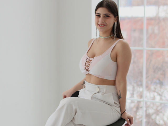 MaryGibbs with medium tits, wearing a pink lace-up crop top and white pants, sitting on a black chair, smiling, brown hair, choker, tattoo on right arm, daylight.