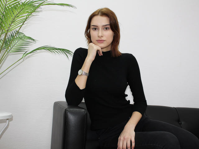 AmandaGate, with brown hair, black long-sleeve shirt, and black pants, sits on a black leather couch, hand on chin, wearing a silver watch, next to a green plant.