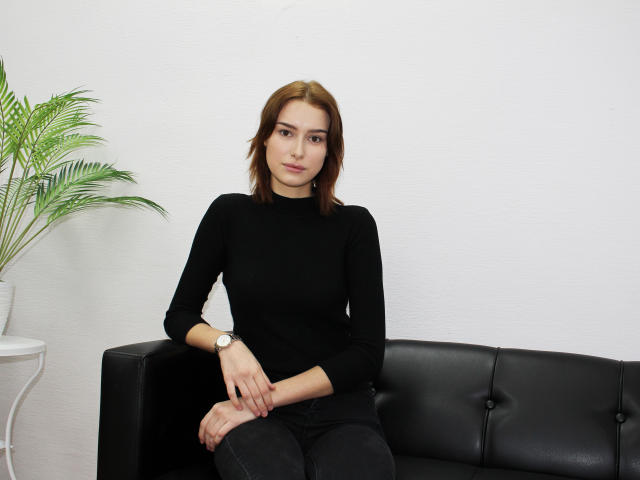 AmandaGate with brown hair, black turtleneck, and black pants sits on a black leather couch, wearing a watch, next to a white pot with green palm leaves.
