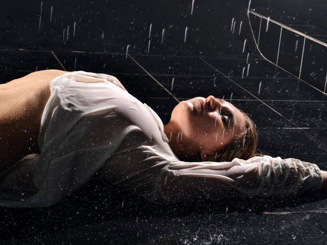 NadinTurner lies on wet black floor, arms above head, wearing soaked white shirt, rain falling on her face, wet brown hair, eyes closed, chest exposed, wet skin glistening.