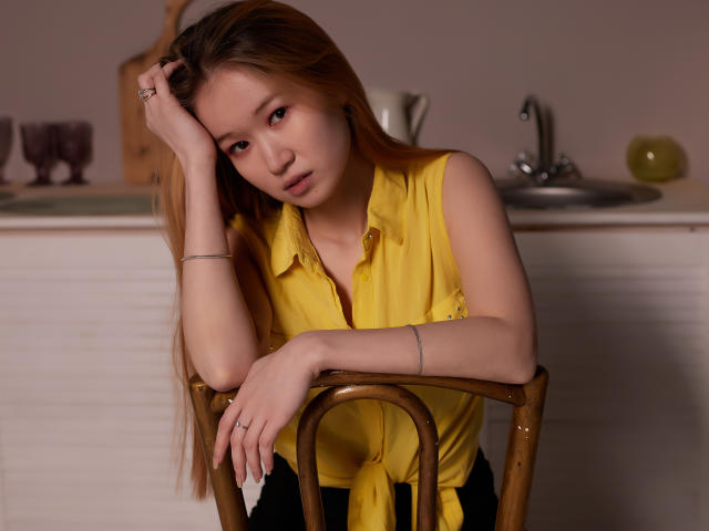 BarbaraWayne with long blonde hair, wearing a yellow sleeveless shirt, leaning on a wooden chair, hand in hair, looking tired, pink eye makeup, silver bracelet, and ring.