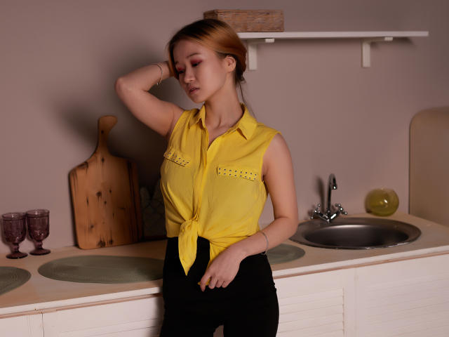 BarbaraWayne with light brown hair, yellow sleeveless shirt tied at the front, black pants, hand in hair, standing in kitchen, wooden board, glasses, and green placemats.