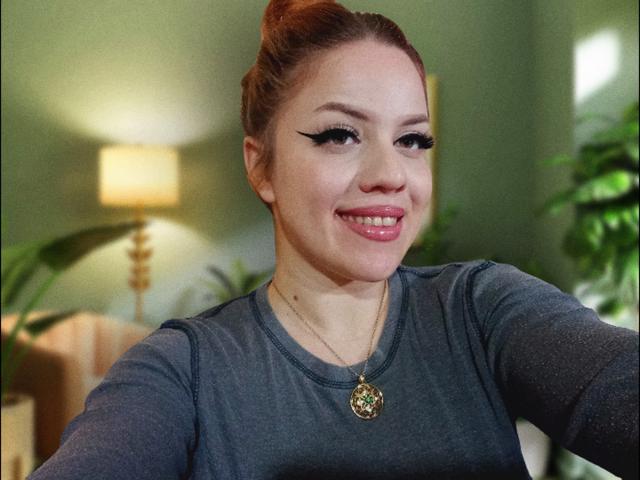 SpicyGrace with red hair, green eyes, and thick black eyeliner, wearing a gray top and gold necklace with a green gem, smiling at the camera.