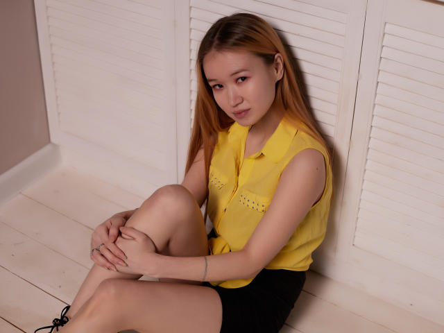 BarbaraWayne sitting on a white floor, wearing a yellow sleeveless shirt and black shorts, legs crossed, arms hugging knees, looking at camera with a slight smile.