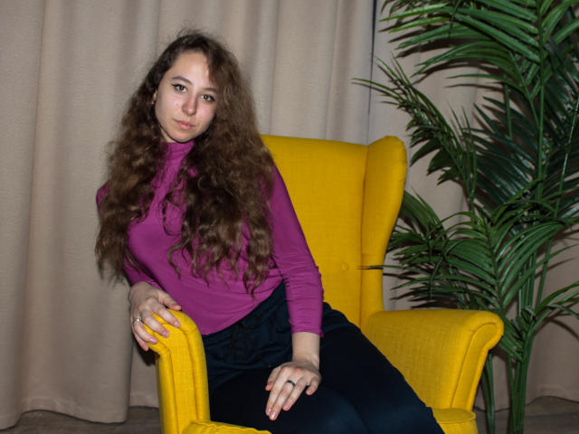 Rachel Thornton mit lockigem, braunem Haar, einem violetten Rollkragenpullover, schwarzen Hosen, einem gelben Sessel und einer grünen Pflanze, sitzt mit einem entspannten, leicht verspielten Ausdruck und zeigt ihre niedlichen, kleinen Brüste..