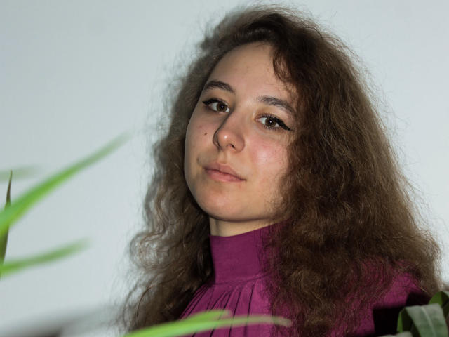 Rachel Thornton heeft krullend bruin haar, een neusring en draagt een paars top met een hoge halslijn. Haar bruine ogen zijn iets omhoog getrokken en ze kijkt naar de camera..