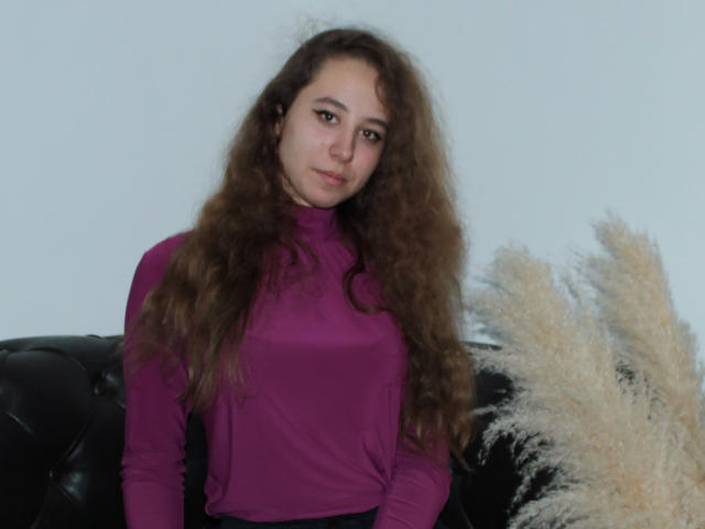 Rachel Thornton mit langen, lockigen, braunen Haaren, die ein violettes Rollkragenpullover trägt, sitzt auf einer schwarzen, mit Leder bezogenen Couch. Rechts daneben befinden sich Büschel von beigem Pampasgras..
