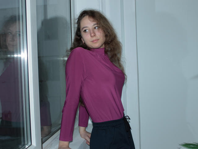 Rachel Thornton, avec ses cheveux bruns ondulés, portant un haut violet à manches longues et un pantalon noir, appuyée contre une fenêtre, avec un reflet visible, regardant légèrement sur le côté, expression neutre..