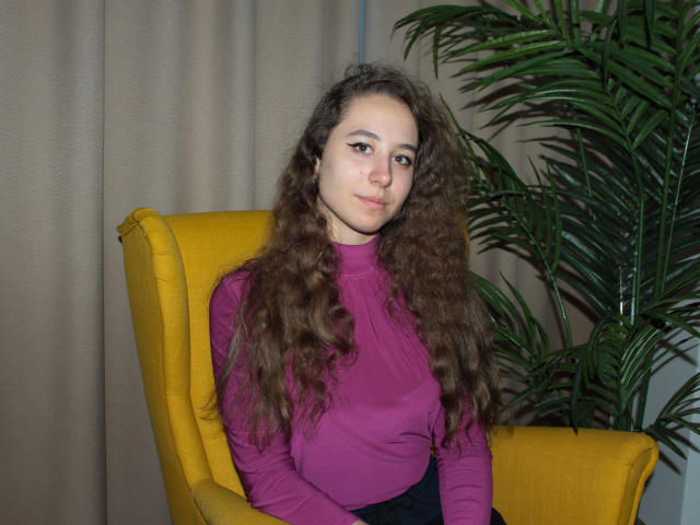 Rachel Thornton, con cabello castaño rizado y largo, una blusa morada de manga larga, una silla amarilla y una planta verde en maceta, sentada con una ligera sonrisa, mirando a la cámara..