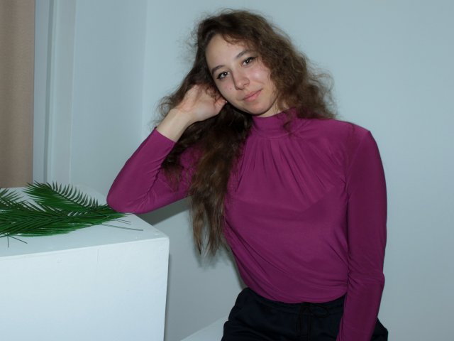 Rachel Thornton mit lockigem, braunem Haar, die ein langes, violettes Oberteil, schwarze Hosen und ein leichtes Lächeln trägt, berührt ihren Kopf mit ihrer rechten Hand und steht neben einem weißen Karton..