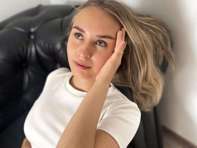 MargoWard with light brown hair, green eyes, and a white shirt, touching her face with a hand, sitting on a black leather chair, looking up with a slight smile.