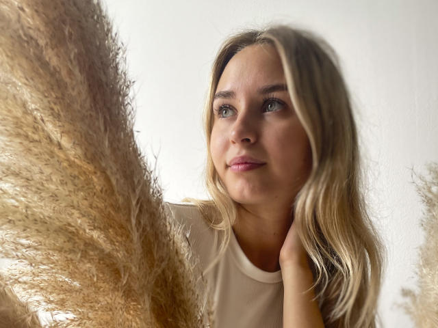 MargoWard with blonde hair, blue eyes, and pink lips, looking up at large, fluffy, beige puffs, wearing a white shirt, hand touching neck.