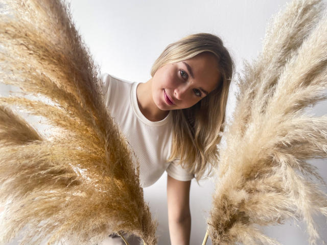 MargoWard with blonde hair, white t-shirt, and a smile, holding large puffy tan grasses, leaning forward, eyes looking at camera, soft natural light.