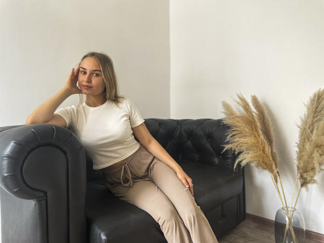 MargoWard with blonde hair, white tee, and beige pants, lounging on a black leather sofa, hand on cheek, pensive expression, pampas grass in a vase to the right.
