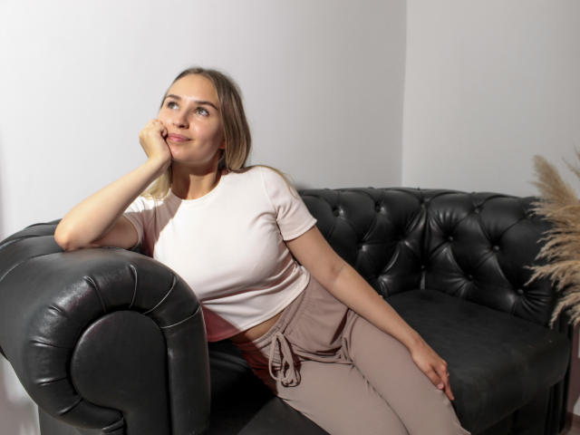 MargoWard with blonde hair, white top, and beige pants, sitting on black leather couch, hand on cheek, looking up, smiling, soft lighting, casual, relaxed, cute.