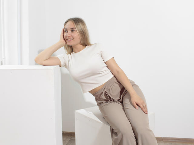 MargoWard with blonde hair, white t-shirt, and beige pants, leaning on a white cube, smiling, showing small tits and a cute ass.