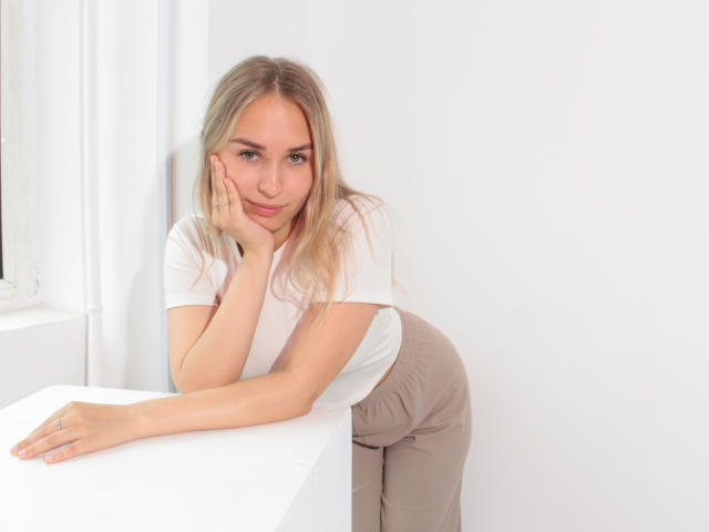 MargoWard with blonde hair, white tee, and beige pants, leaning on a white table, hand on cheek, subtle smile, blue eyes, soft light, casual, sexy, cute.