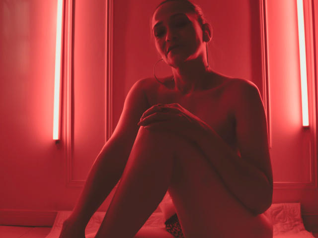 AgathaStone is nude, sitting with knees up, hands on knees, red neon light casting glow, small tits, subtle ass, her face shadowed, ears with hoop earrings, looking forward.