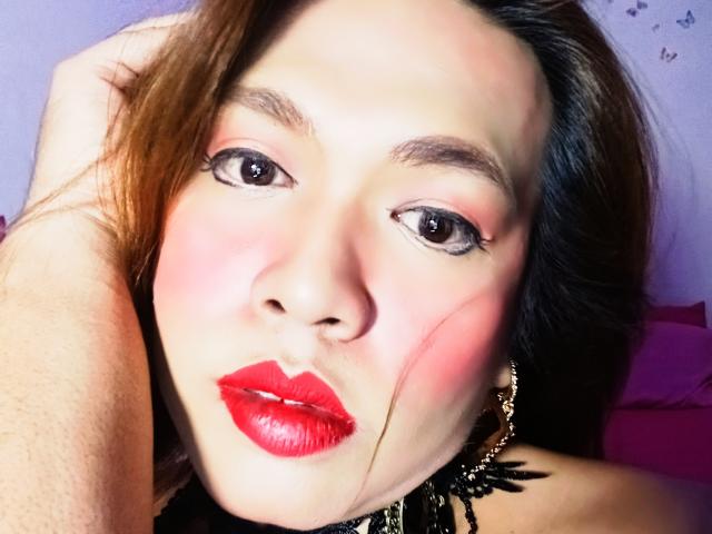 JadeWithLove has bright red lips, pink blushed cheeks, black eyeliner, and brown hair, with a hand in her hair and a black, leafy earring. Her eyes are slightly open.