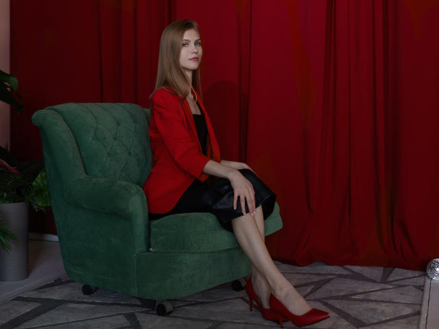 PhoebeDyson sits on a green chair, wearing a red jacket, black dress, and red heels, with her ass slightly raised, looking serious, and her pussy not visible.