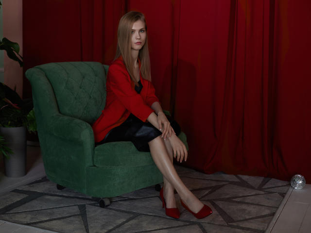 PhoebeDyson with blonde hair, red jacket, black skirt, shiny legs, red heels, sitting on green chair, red curtains, plant on left, silver ball on floor.