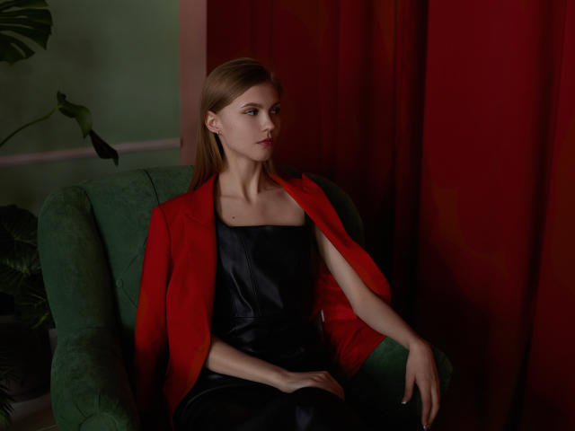 PhoebeDyson with straight brown hair, wearing a red blazer over a black leather dress, sitting on a green armchair, looking slightly bored, with red curtains in the background.