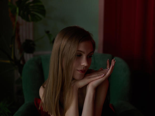 PhoebeDyson with long blonde hair, hands resting on her chin, sitting in a green chair, looking down with a slight smile, red curtains in the background, soft lighting.