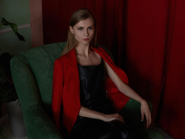 PhoebeDyson with long blonde hair, red blazer, black leather dress, sitting on green chair, red curtain background, looking serious, hands resting on chair. Boobs visible under dress.