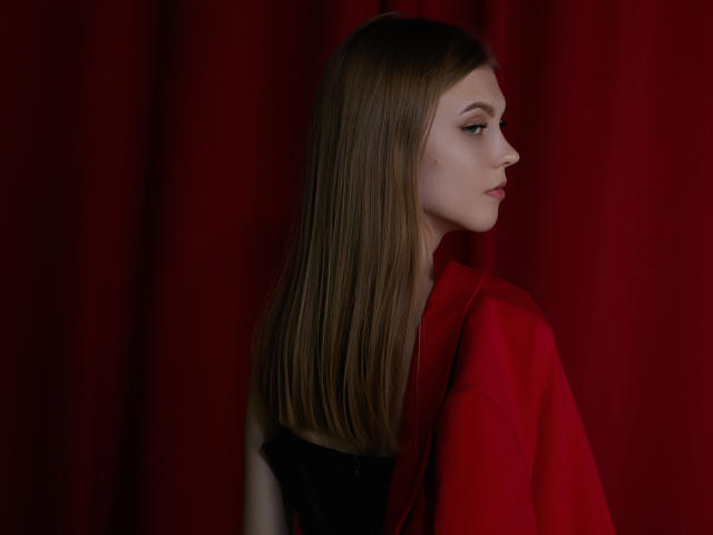 PhoebeDyson with long brown hair, wearing a red jacket, black top, side profile, red curtain background, looking right, subtle makeup, calm expression, slight freckle on cheek.