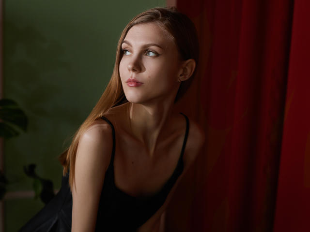 PhoebeDyson has light skin, long brown hair, and is wearing a black dress. She's looking to the side with a slight smile, standing near red curtains.