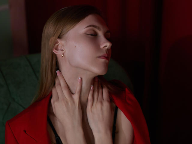 PhoebeDyson with eyes closed, wearing a red jacket, black lingerie, hands on neck, long blonde hair, soft pink nails, slight pink blush, calm expression, red background.