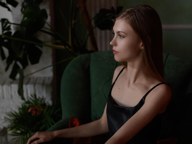 PhoebeDyson with long brown hair, small tits, wearing a black satin dress, sitting in a green chair, looking to the side, soft lighting, subtle red lipstick, earrings.