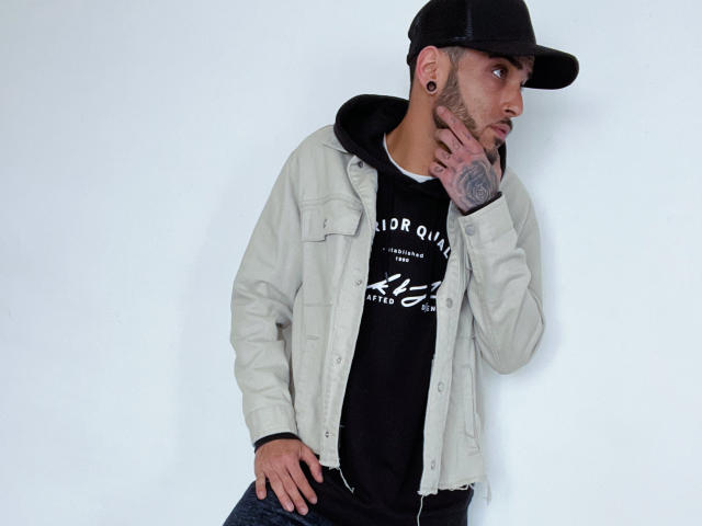 HardyGeorge, wearing a black cap, white jacket, black hoodie, and blue jeans, touches his tattooed cheek with his left hand, looking to the side with a serious expression.