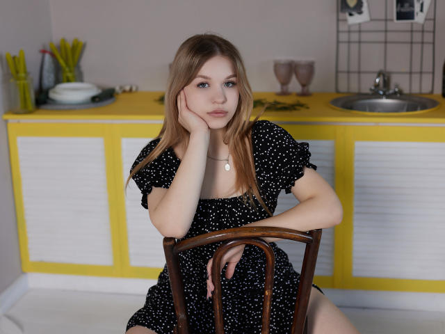 SimonaForman with blonde hair, wearing a black polka dot dress, sits on a wooden chair, resting her head on her hand, showing her small tits and cute face.