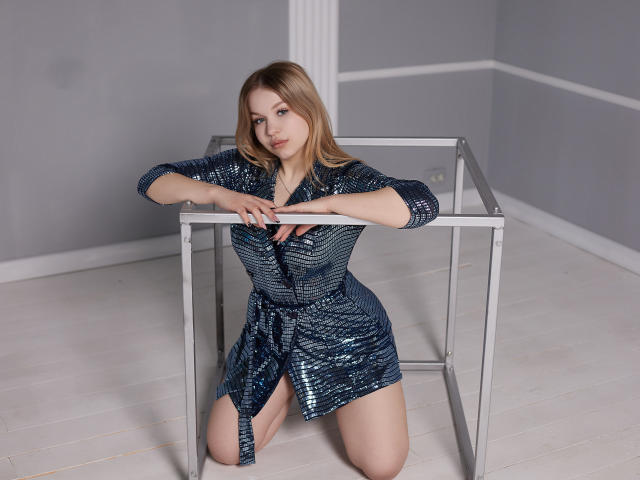 SimonaForman kneeling, wearing a blue sequined dress, arms resting on a metal frame, legs apart, showing her pussy, with a neutral expression, in a minimalist room.