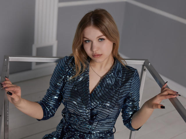 SimonaForman with blonde hair, wearing a blue sequined dress, sitting on a metal chair, hands gripping the frame, looking at the camera, black nail polish, neutral expression.