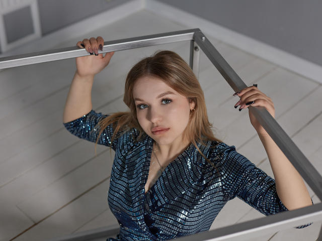 SimonaForman with blue sequined dress, holding metal railing, blonde hair, green eyes, and black nail polish, standing on white wooden stairs, looking up at camera.