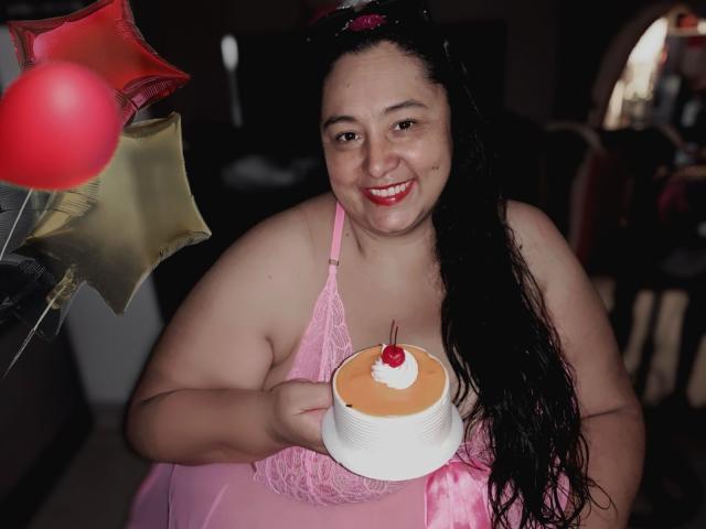 TistMature, a chubby woman with long black hair, pink lace lingerie, holding a small cake with a cherry, smiling at the camera. Balloons in the background.